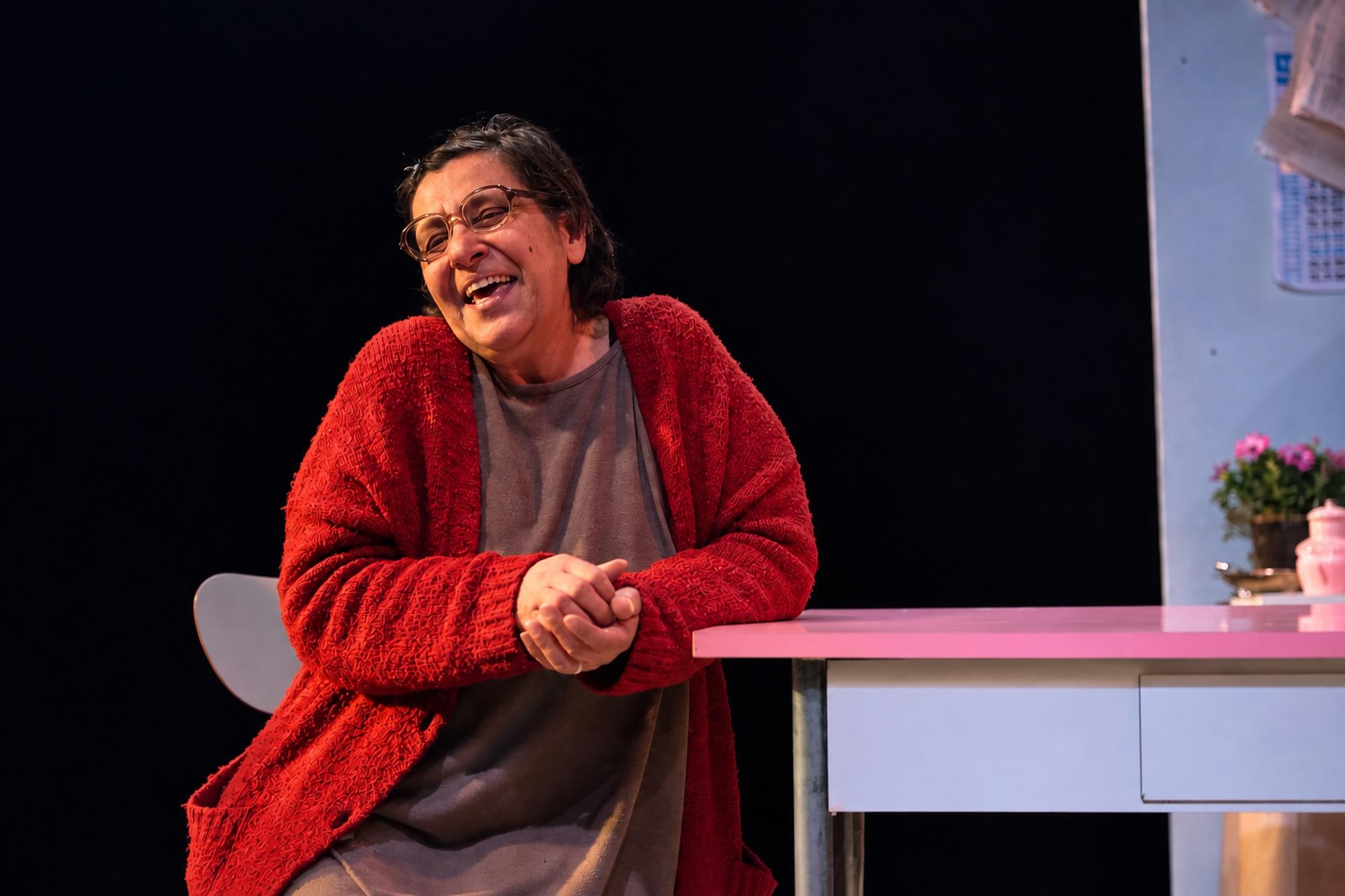 Una donna sorridente in cardigan rosso, appoggiata con le braccia a un tavolo rosa.