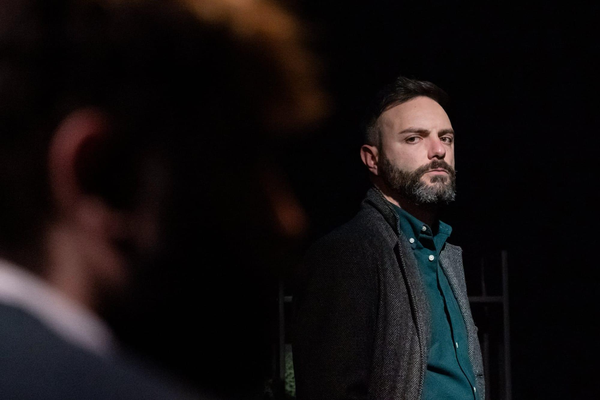Un uomo con la barba in scena davanti a un altro interprete sfocato.