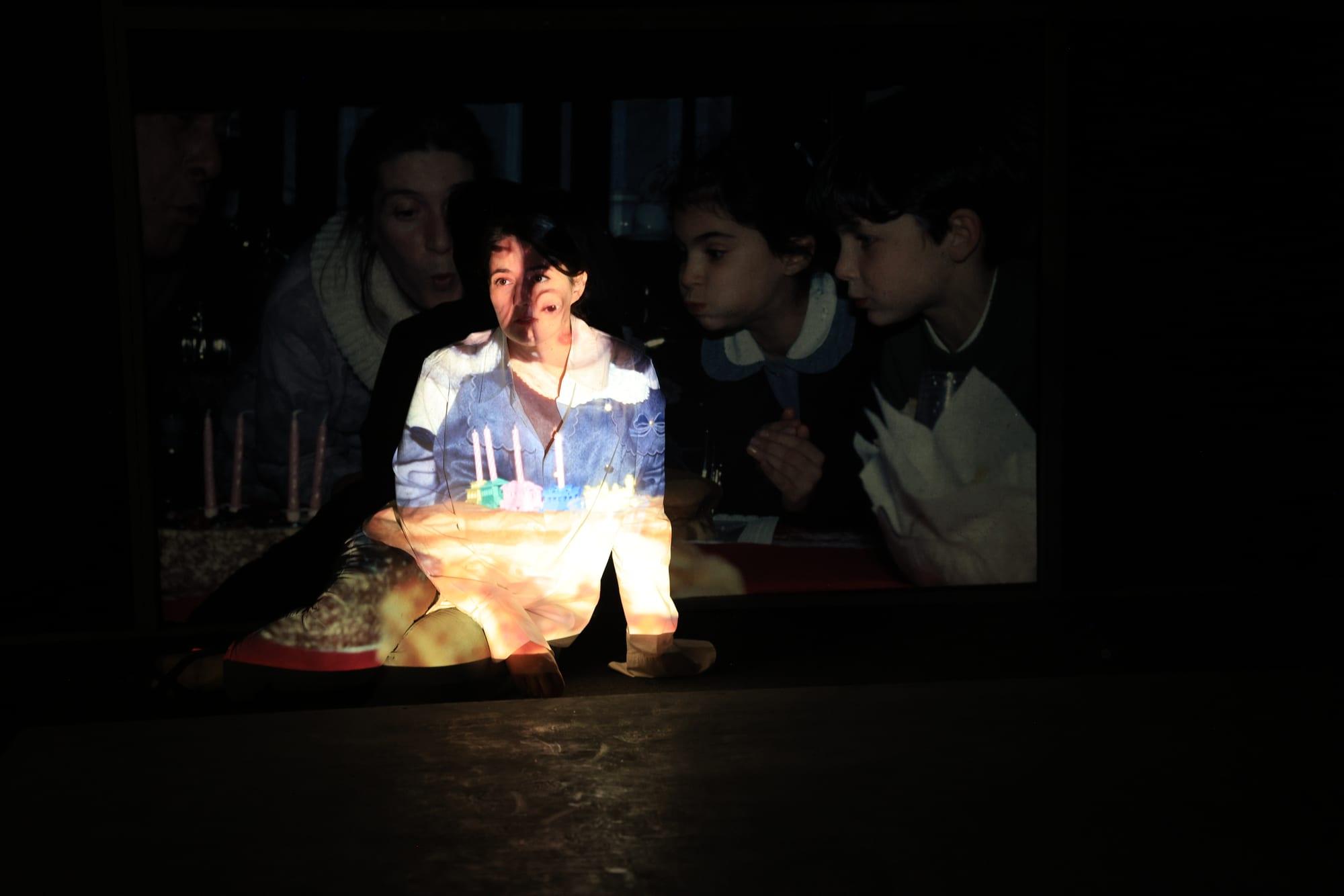 Una donna seduta sul palco, attraversata dalla proiezione di una scena di compleanno.