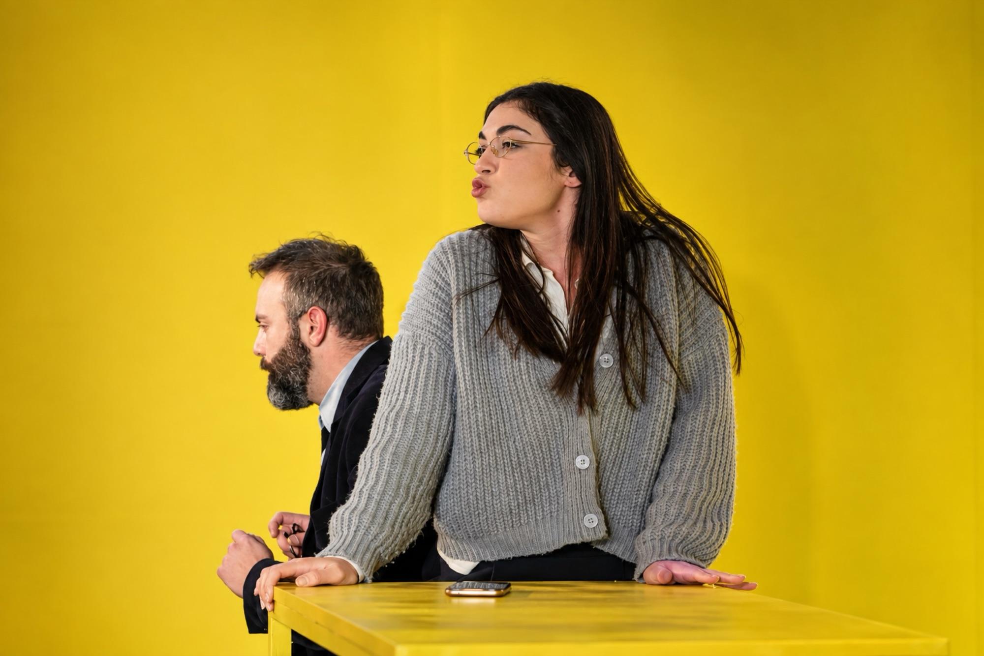 Una donna in cardigan grigio parla dietro a un tavolo giallo, alle sue spalle un uomo seduto guarda altrove.